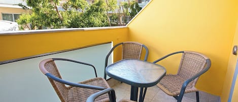 A balcony with wicker chairs and a table, overlooking a yellow building and trees.