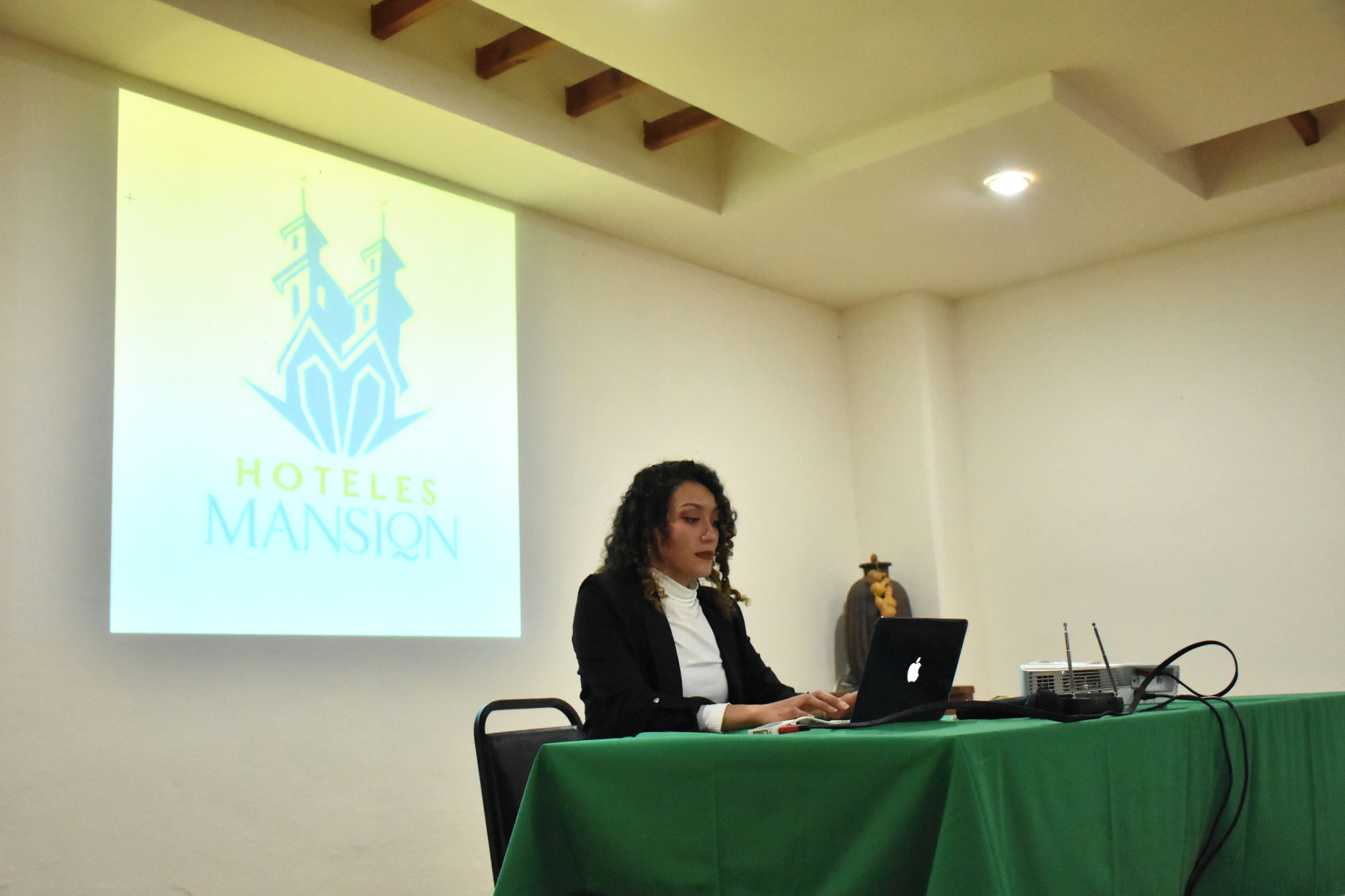 Una presentación con una proyección del logotipo HOTELES MANSION, una computadora portátil y un proyector sobre una mesa.
