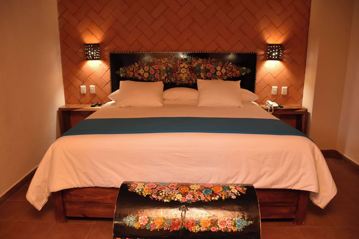 Una cama bien hecha con dosel floral, mesitas de noche con lámparas y un banco con motivos florales.