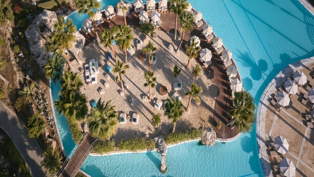 Eine Vogelperspektive zeigt einen luxuriösen Urlaubsort mit einem zentralen Pool, umgeben von Palmen, Liegestühlen und Sonnenschirmen.