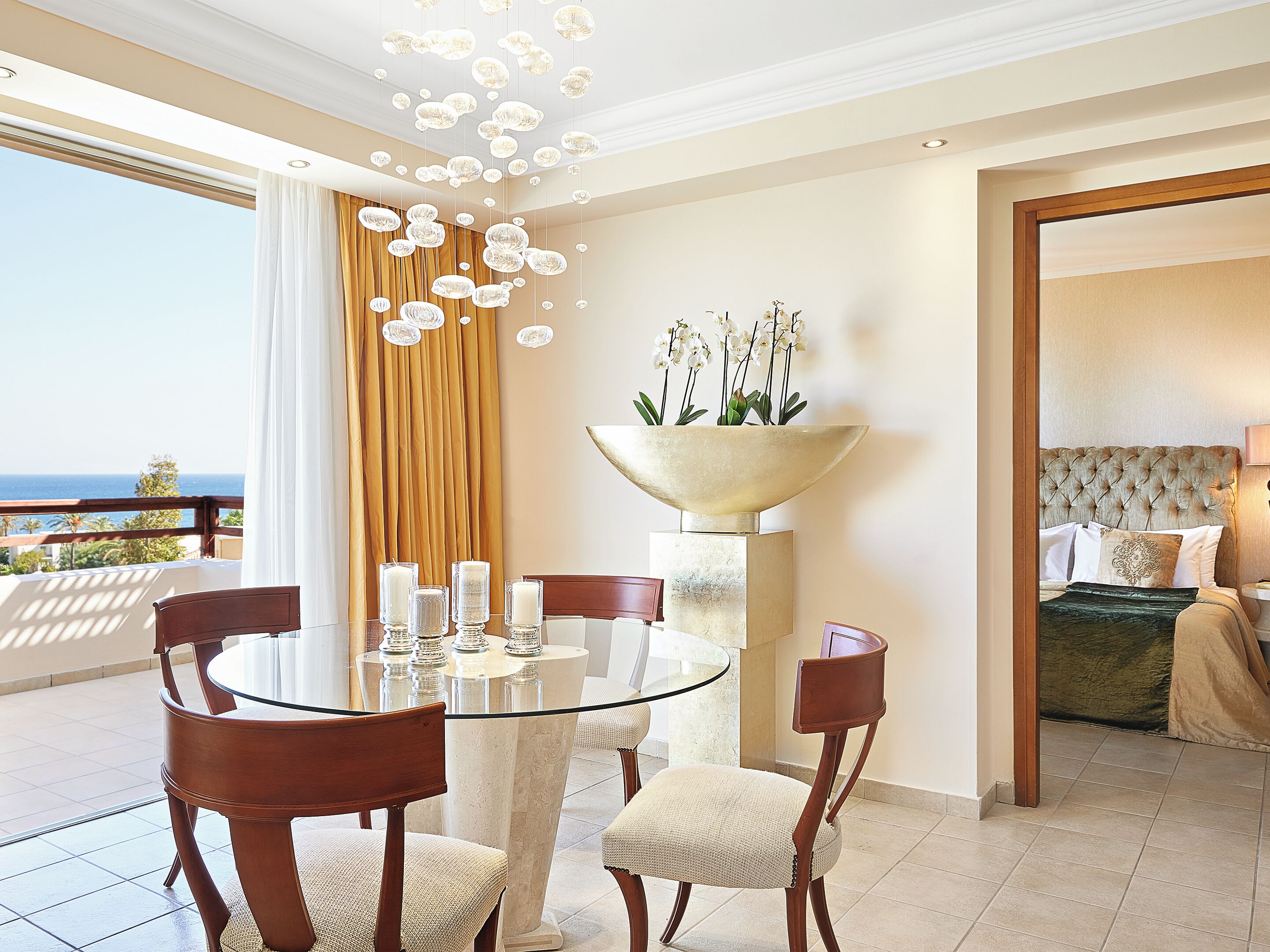 A hotel room with a balcony, a round glass table with chairs, a bed with a tufted headboard, and a decorative bowl with flowers.
