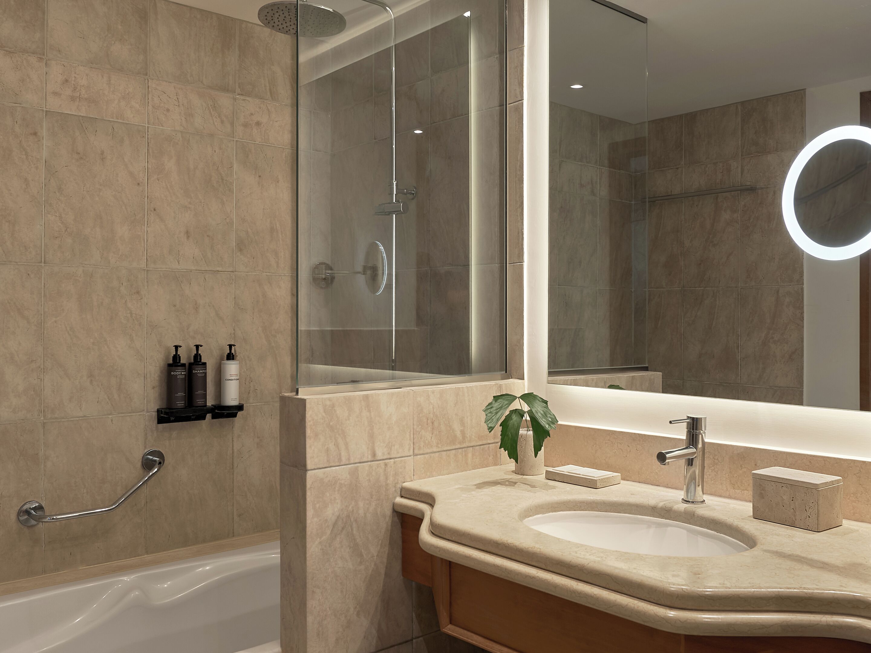 Un baño moderno con ducha de vidrio, tocador con lavabo y un espejo grande con luz circular.