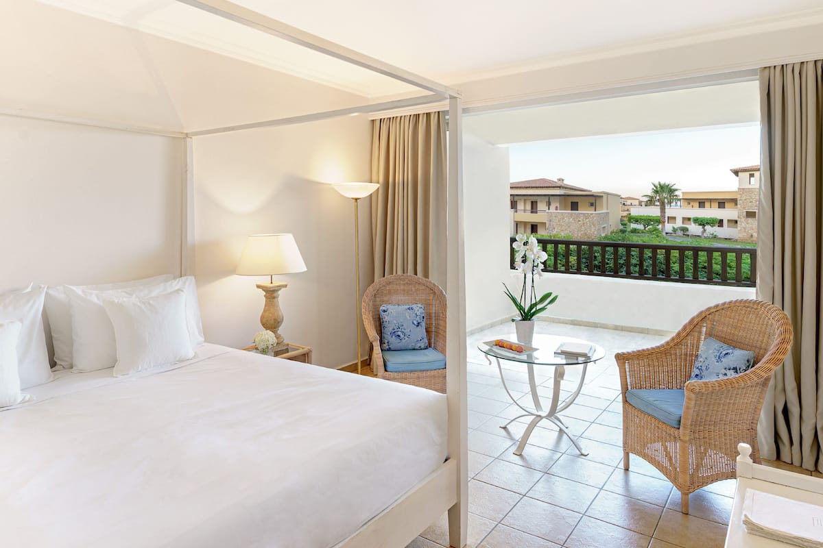 Hotelkamer met een groot bed, nachtkastje met lamp, twee rieten stoelen, een klein glazen tafeltje met een potorkidee, en een balkon met uitzicht op andere gebouwen en groen.