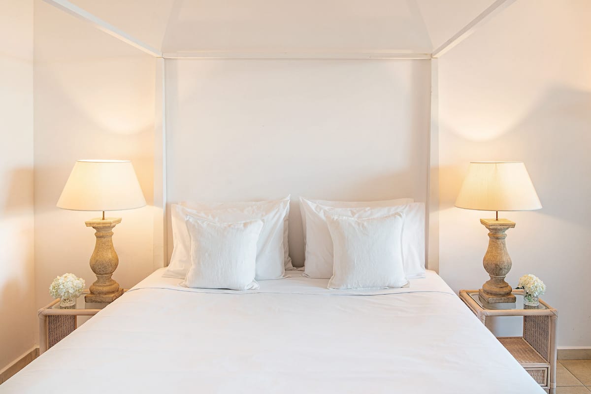 Een netjes opgemaakt bed met witte lakens, twee bedlampen en op elk nachtkastje een klein vaasje met bloemen.