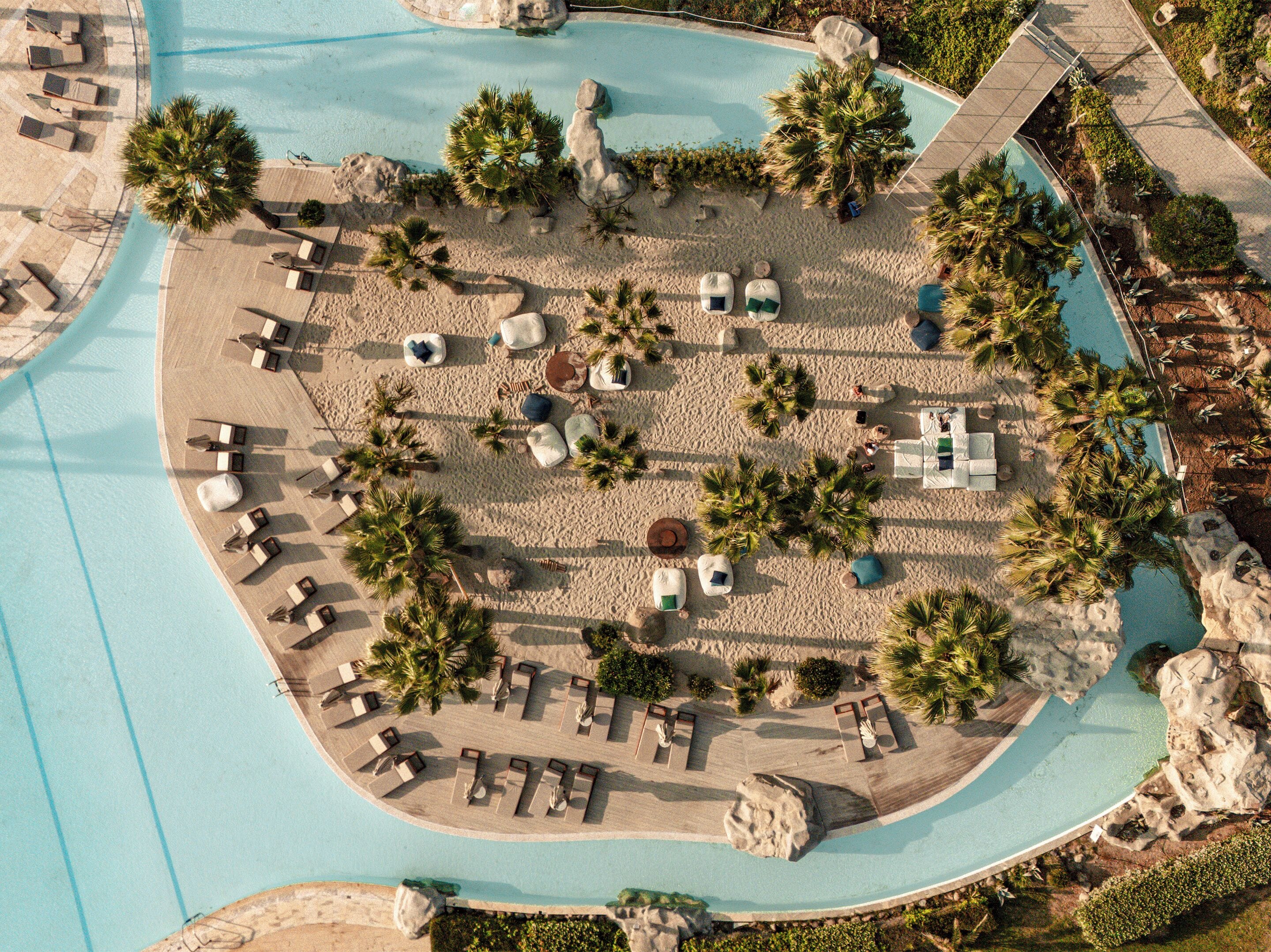 An aerial view of a resort with a pool, sun loungers, and palm trees.