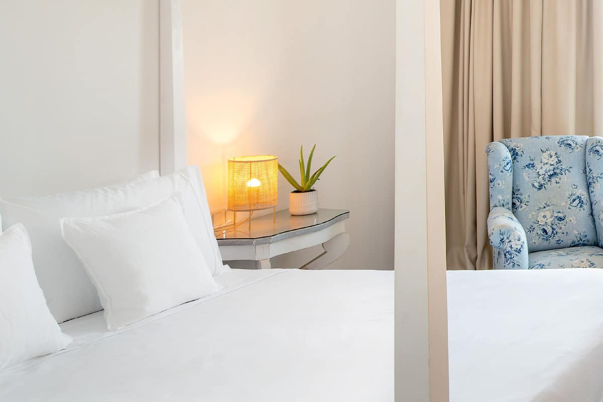 Een hotelkamer met een bed, een nachtkastje met een lamp en een potplant, en een blauwe fauteuil met bloemenprint.
