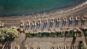 Ein Luftbild eines Strandes mit Reihen von Sonnenschirmen und Liegestühlen, einem gepflasterten Weg und einer felsigen Küstenlinie.