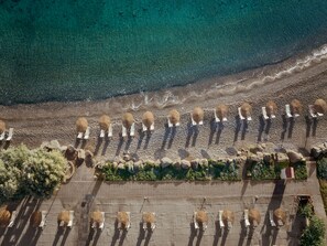 An aerial view of a beach with rows of umbrellas and lounge chairs, a paved walkway, and a rocky shoreline. - Grecotel LUXME Kos (Kos)