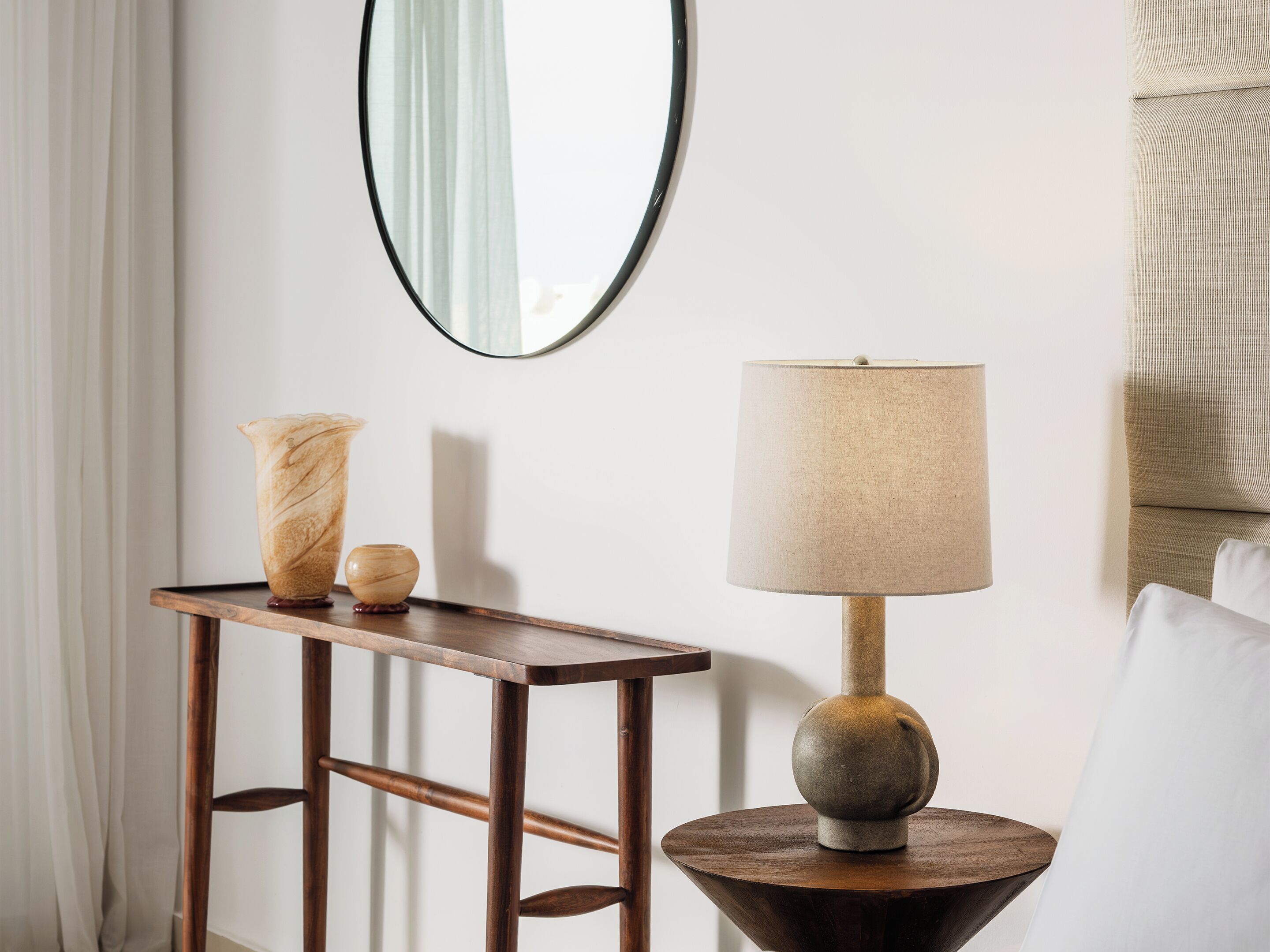 A wooden side table with a lamp and a vase, a mirror on the wall, and a white curtain.
