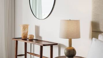 A wooden side table with a lamp and a vase, a mirror on the wall, and a white curtain.