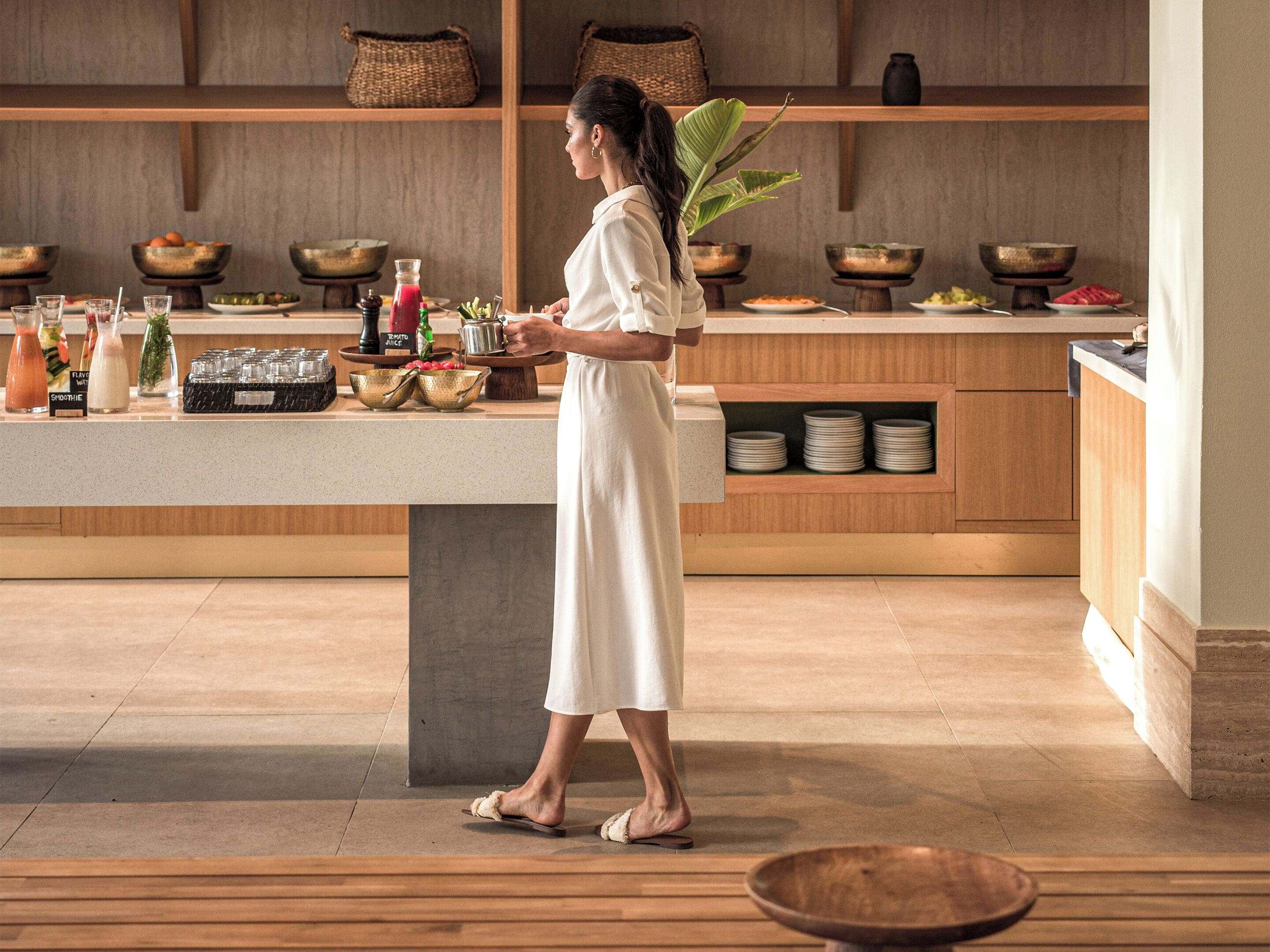 Una mujer con vestido blanco parada en una cocina moderna con estantes de madera, una encimera con varios cuencos y botellas, y una planta grande.