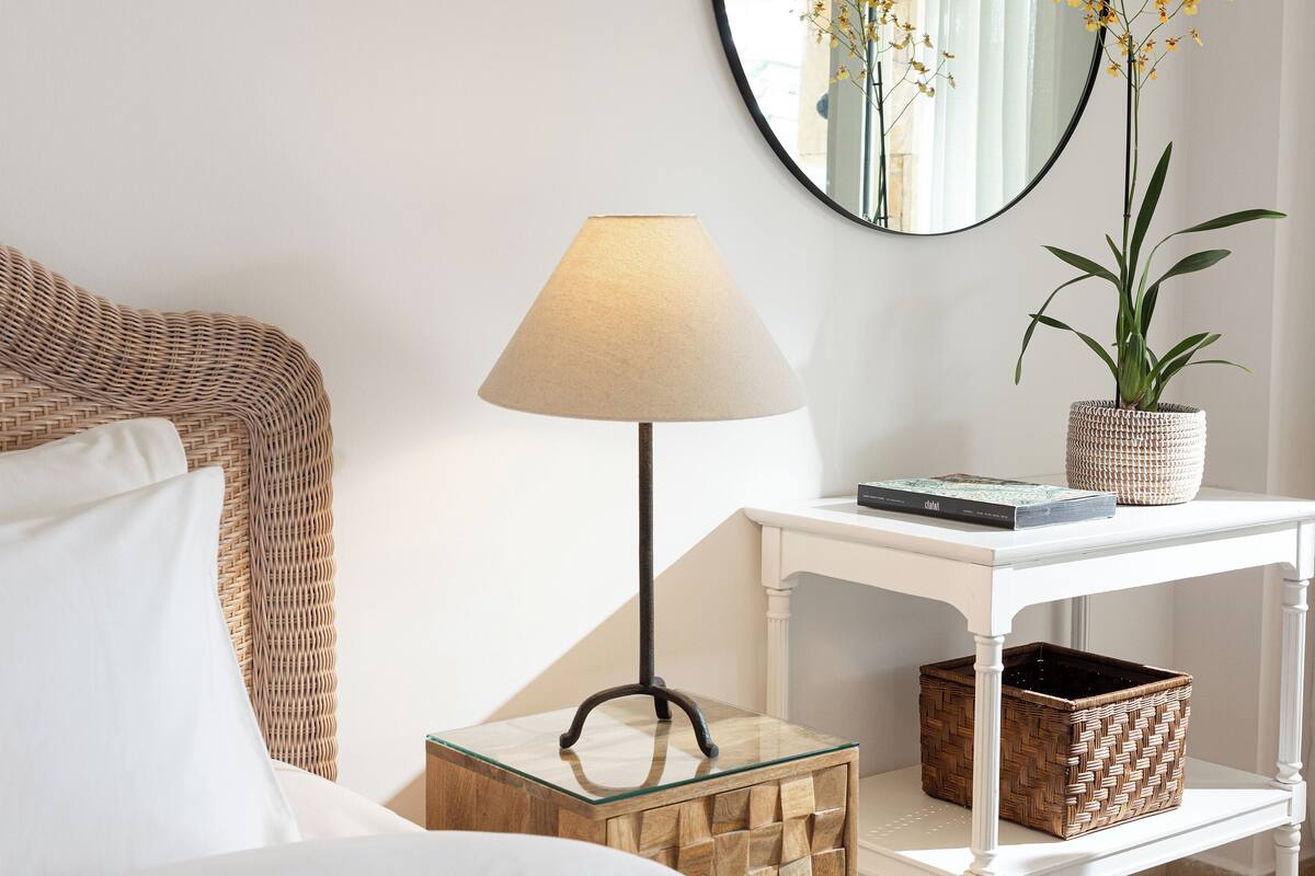 Een keurig opgemaakt slaapkamer met een rieten hoofdbord, een bedlampje, een bloempot, een boek en een geweven mand.
