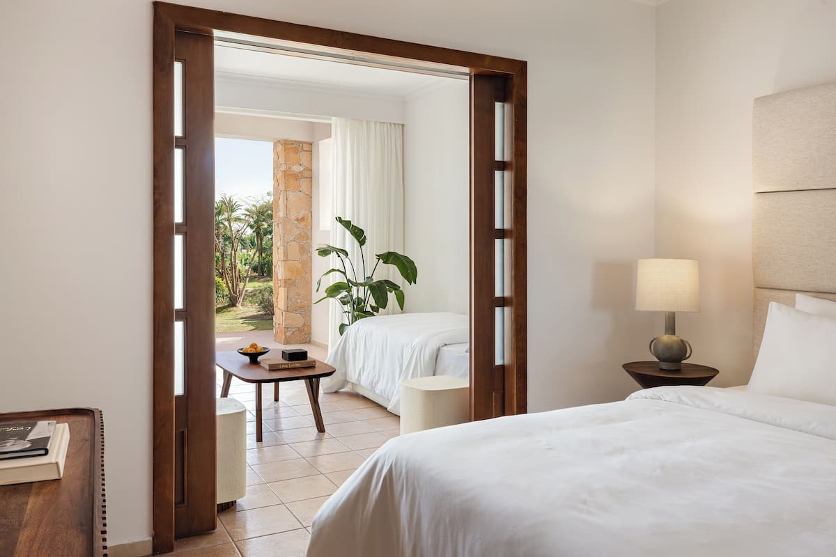 Een hotelkamer met een bed, een nachtkastje met een lamp, een tafeltje met een fruitschaal, en uitzicht op een tuin door een open deur.