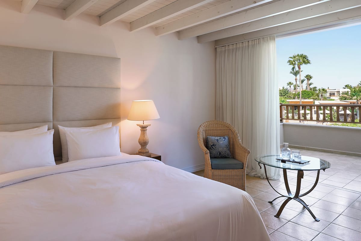 Een hotelkamer met een groot bed, een stoel, een tafel en uitzicht op palmbomen.