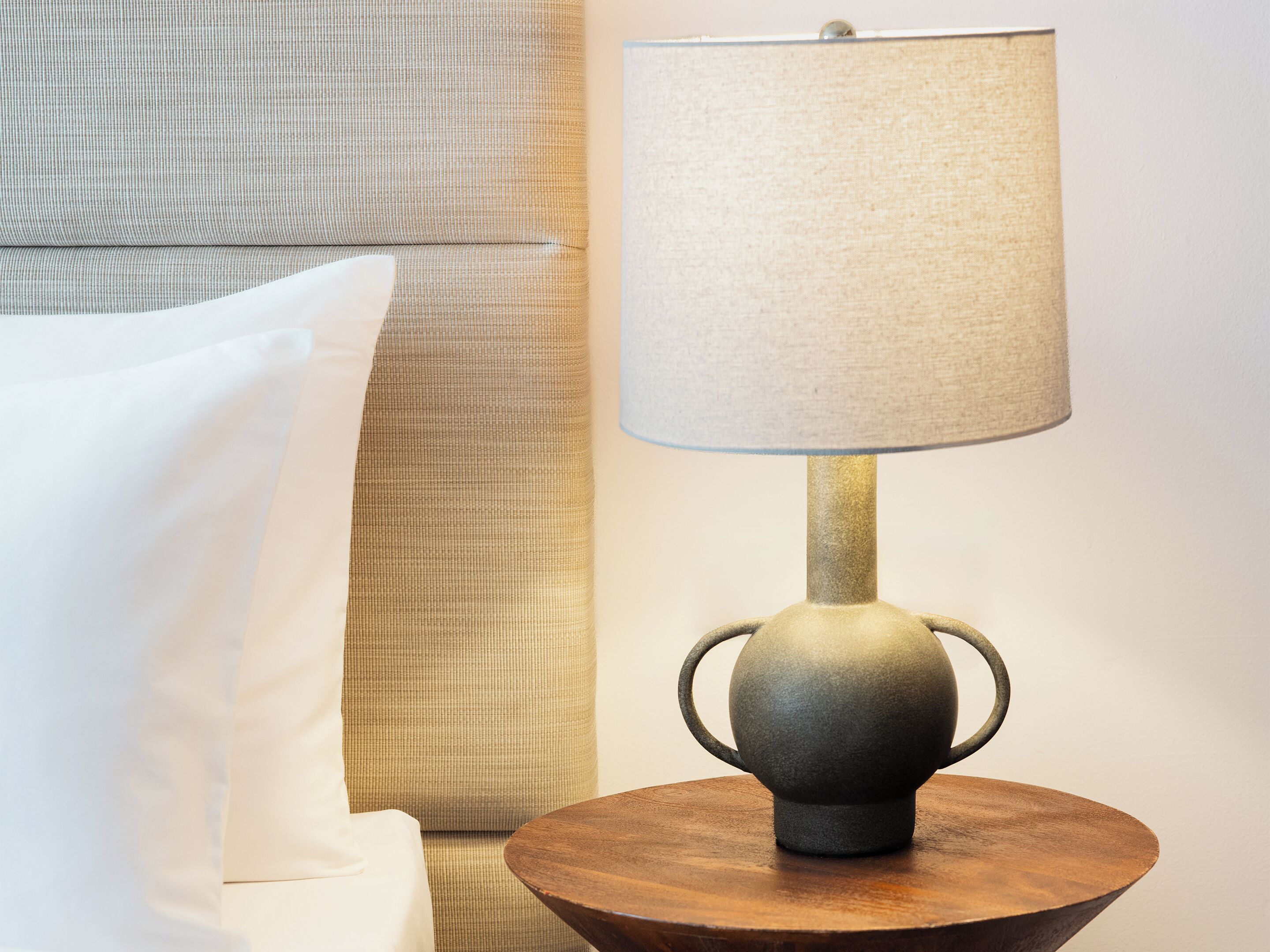 A lamp with a textured shade on a round wooden table, a striped sofa, and a white pillow.