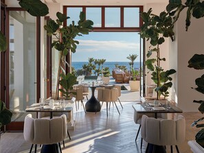 An open-plan dining area with a view of the sea, featuring tables and chairs, potted plants, and a large window. - Grecotel LUXME Kos (Kos)