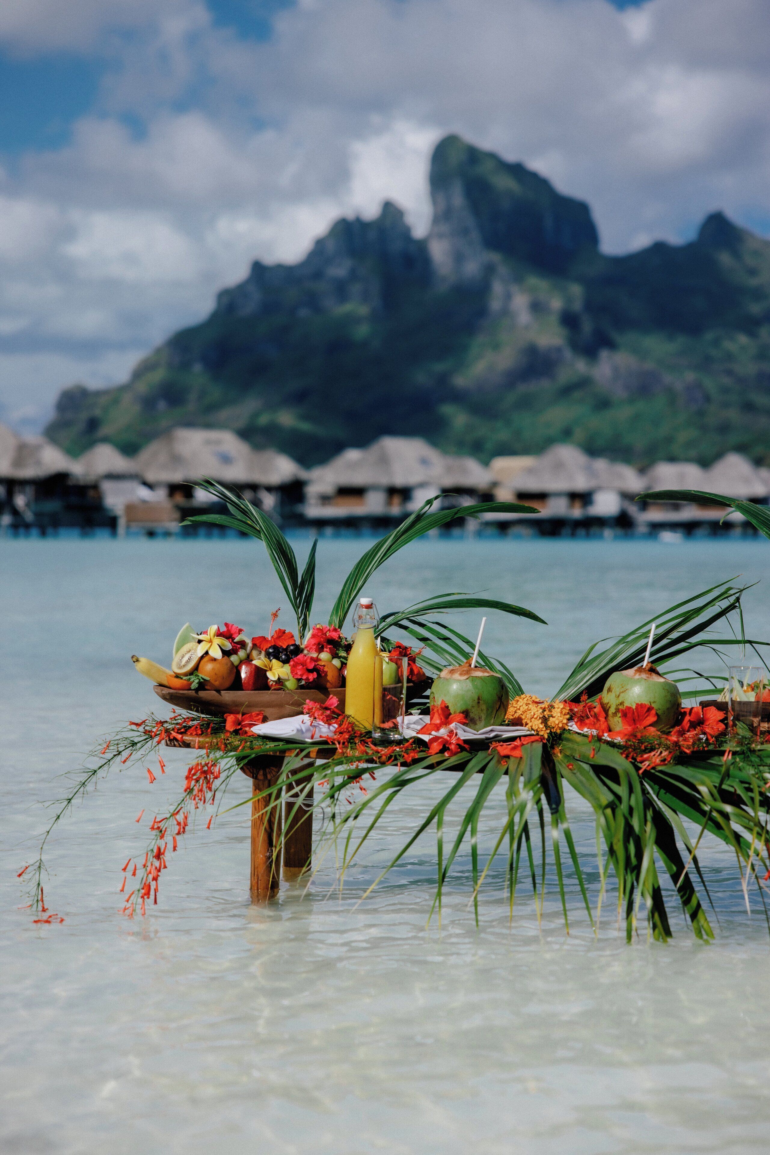A tropical setting with a table set with fruits, flowers, and drinks, overlooking a clear lagoon and overwater bungalows.