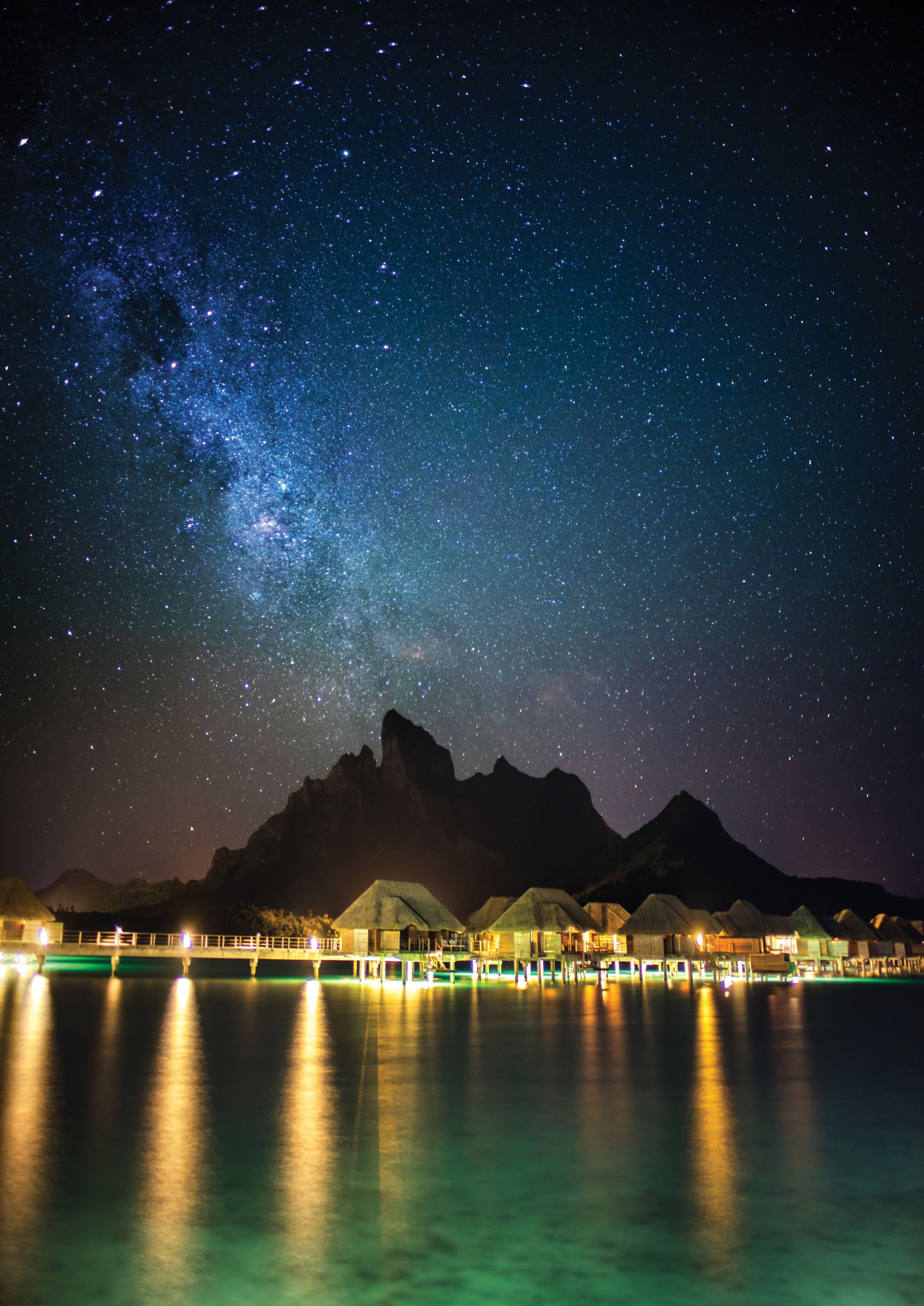 Vista noturna da costa, com céu estrelado, uma cordilheira e um calçadão com edifícios iluminados.