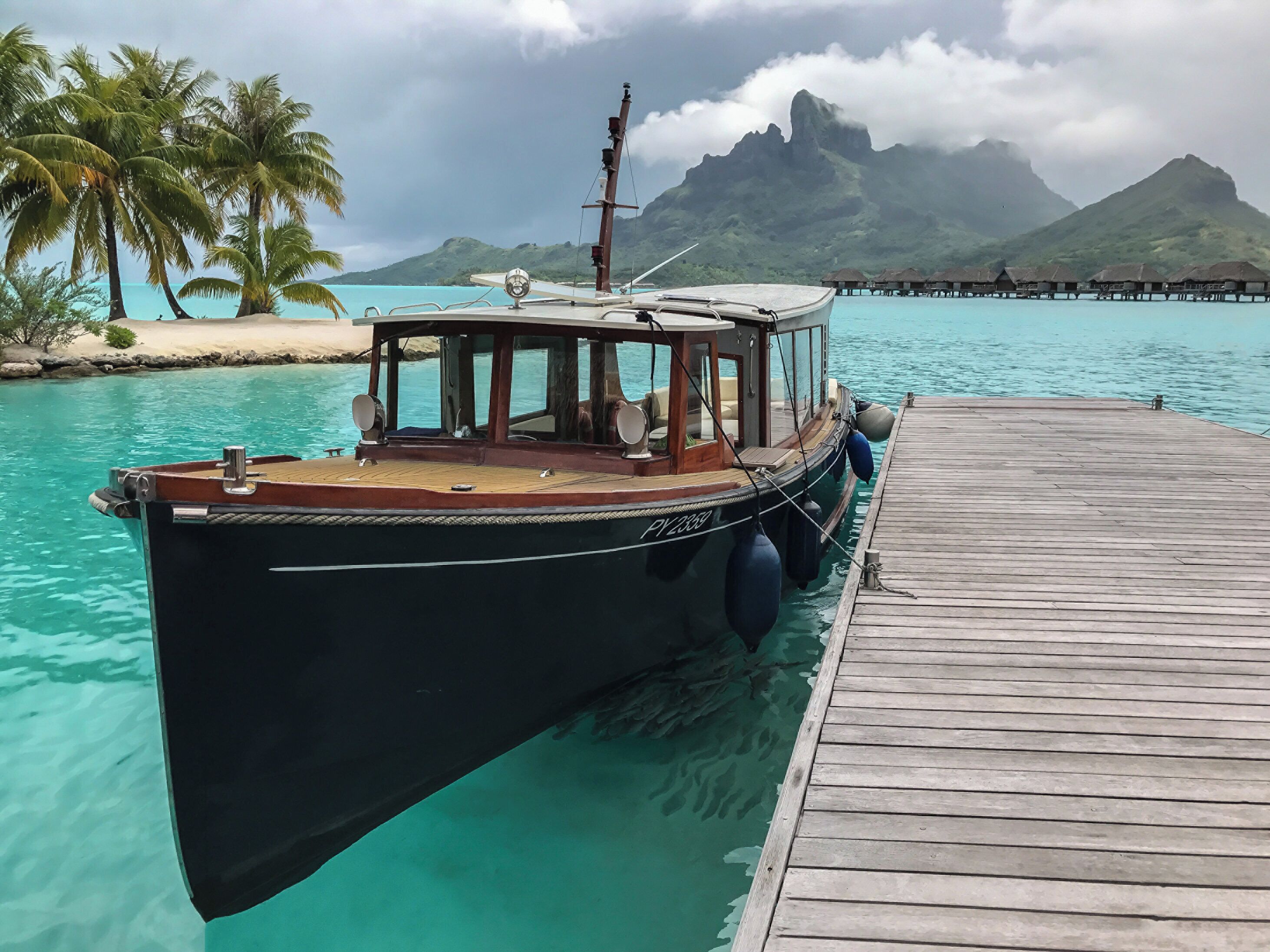 A boat docked at a pier with a wooden deck, surrounded by turquoise water and palm trees.