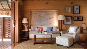 A cozy room with a wooden ceiling, a sofa with cushions, a coffee table with flowers, a chair, and framed pictures on the wall.