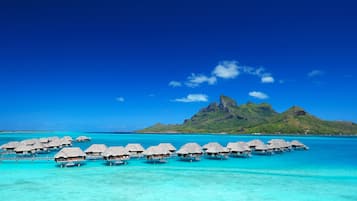 Overwater bungalows with thatched roofs in turquoise waters, a clear sky, and a mountain in the background.