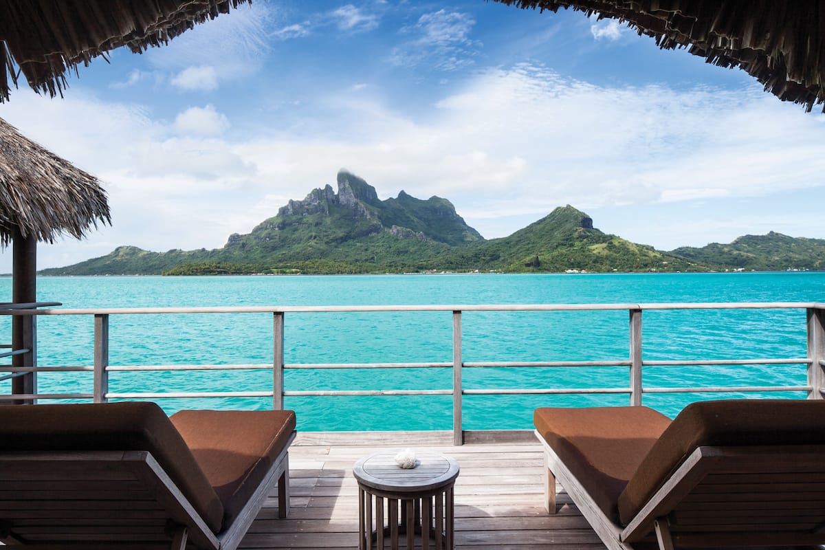 A wooden deck with two lounge chairs and a small table, overlooking turquoise waters and a mountainous landscape.