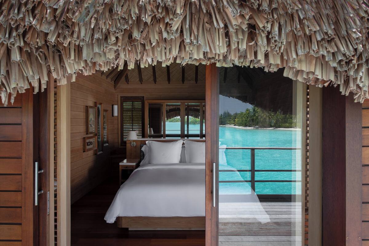 A wooden room with a bed, a nightstand, and a window overlooking a turquoise ocean.