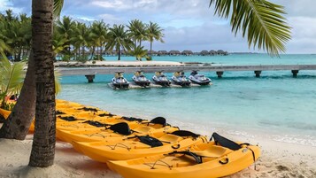Una playa con kayaks amarillos, una zona para motos acuáticas y bungalows sobre el agua al fondo.