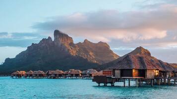Bungalows über dem Wasser mit Reetdächern auf Stelzen, ein klares türkisblaues Meer und eine gebirgige Kulisse.