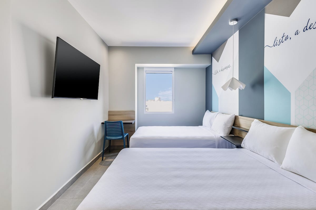 Ein Hotelzimmer mit zwei Betten, einem an der Wand befestigten Flachbildfernseher, einem Fenster mit Blick auf Gebäude und einer Wand mit geometrischem Muster und Text.