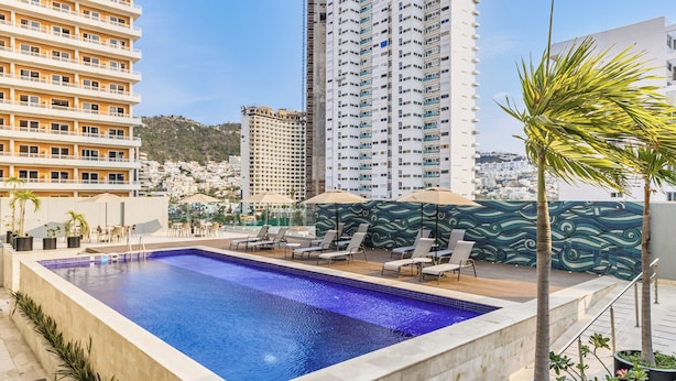 A rooftop pool with lounge chairs and umbrellas, surrounded by high-rise buildings and a mountainous backdrop. - One Acapulco Costera (Acapulco)