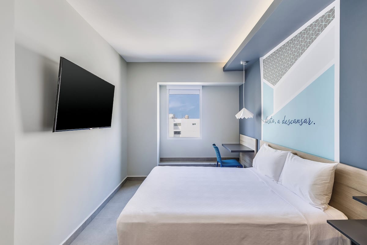 Ein modernes Hotelzimmer mit Bett, Wandfernseher, Schreibtisch und Fenster mit Blick auf die Stadt.