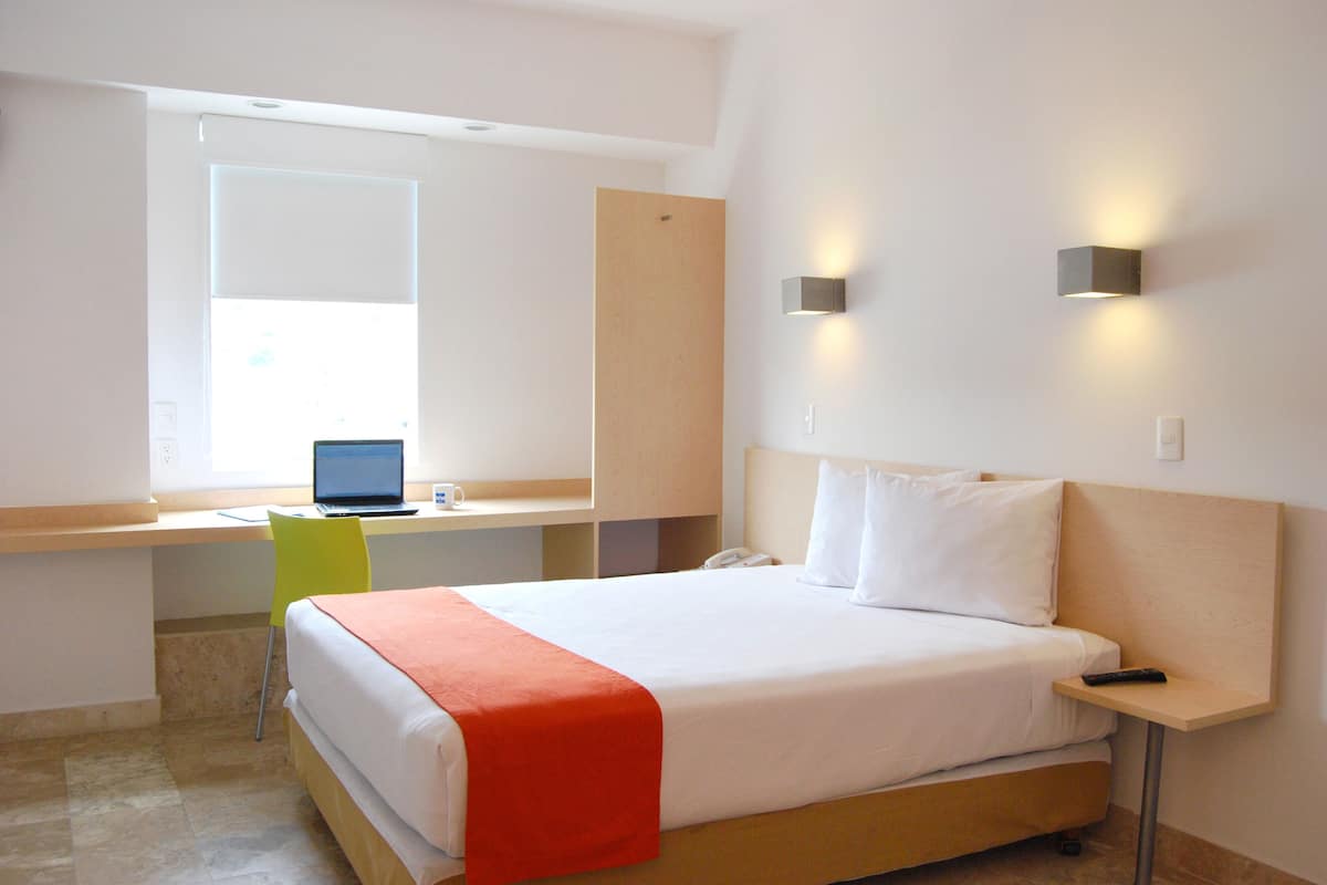 Ein Hotelzimmer mit Bett, Schreibtisch mit Laptop, Stuhl und Nachttisch mit Fernbedienung.
