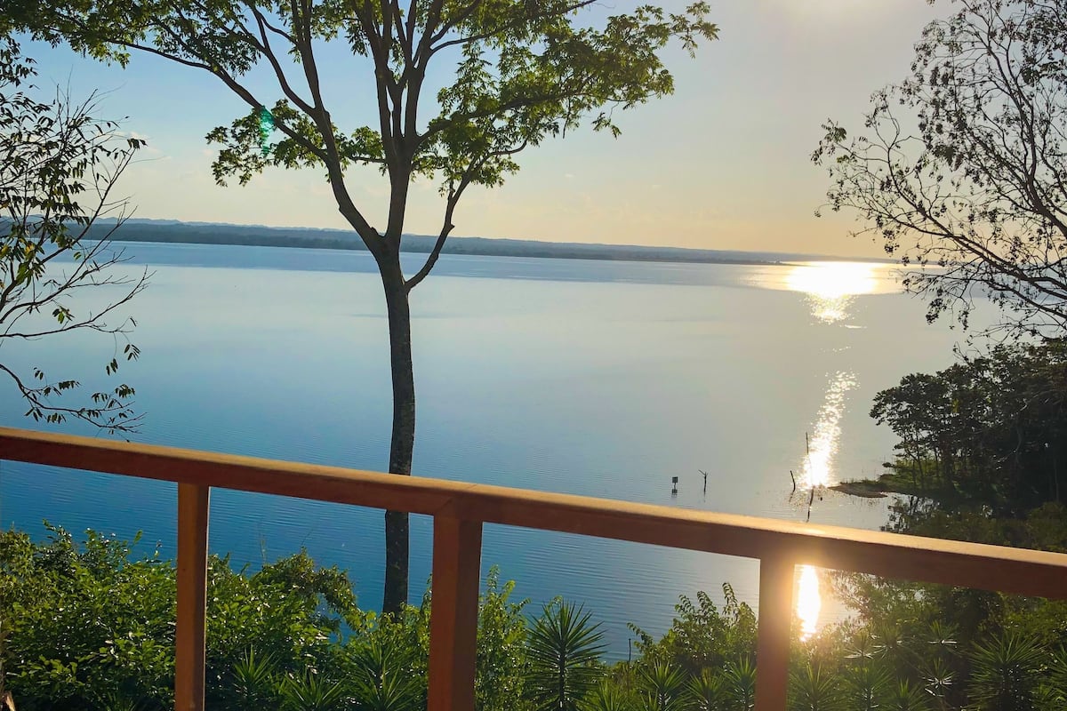 A balcony with a view of a calm lake and a tree in the foreground.