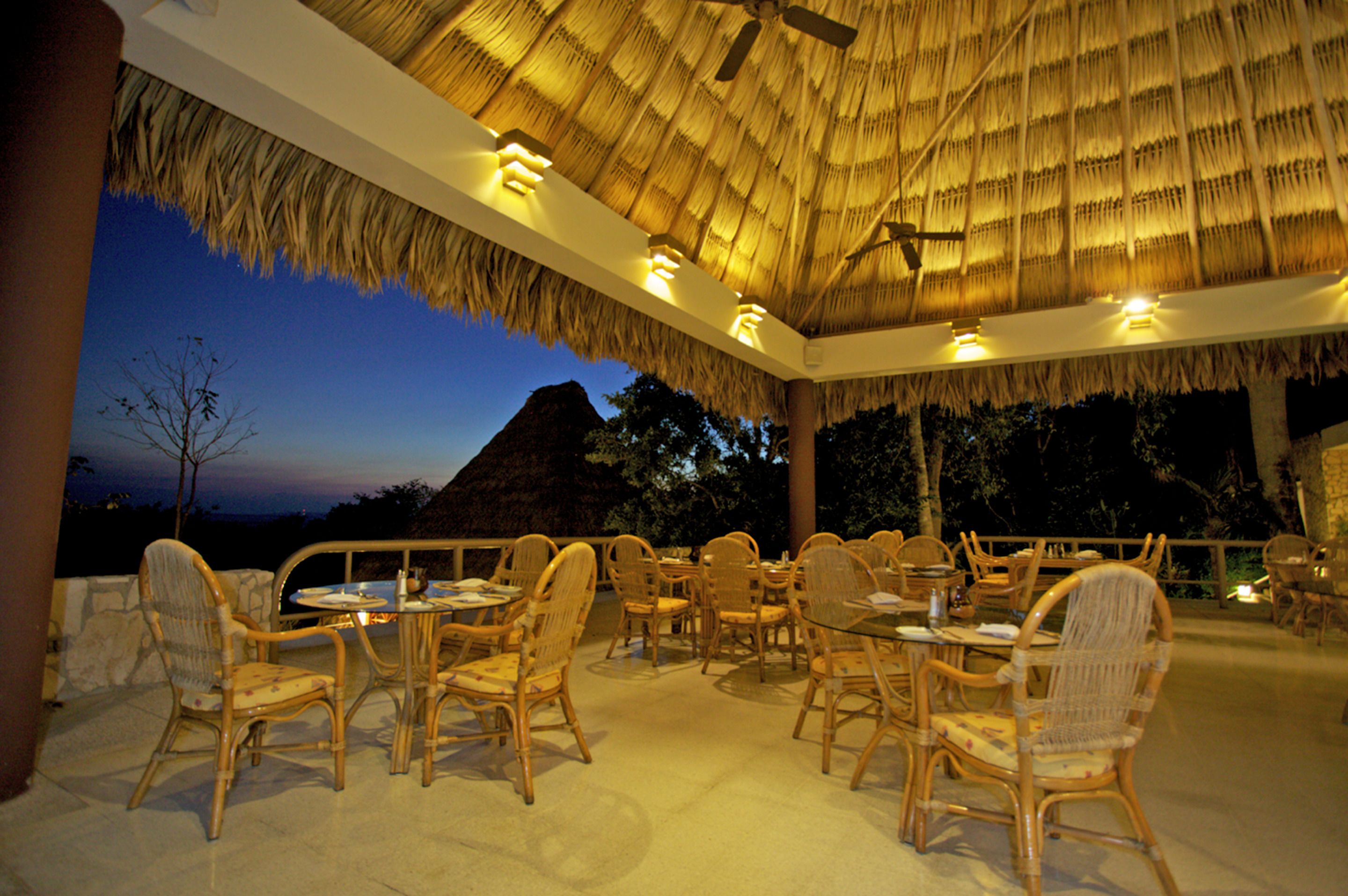 An outdoor dining area with wicker chairs and tables, a thatched roof, and a view of a mountain at dusk.