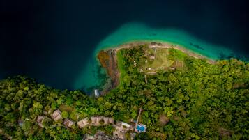 Vista aérea de una propiedad costera con playa, bosque denso y un pequeño grupo de edificios.