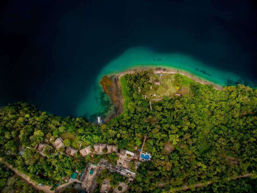 Vista aérea de una propiedad costera con playa, bosque denso y un pequeño grupo de edificios.