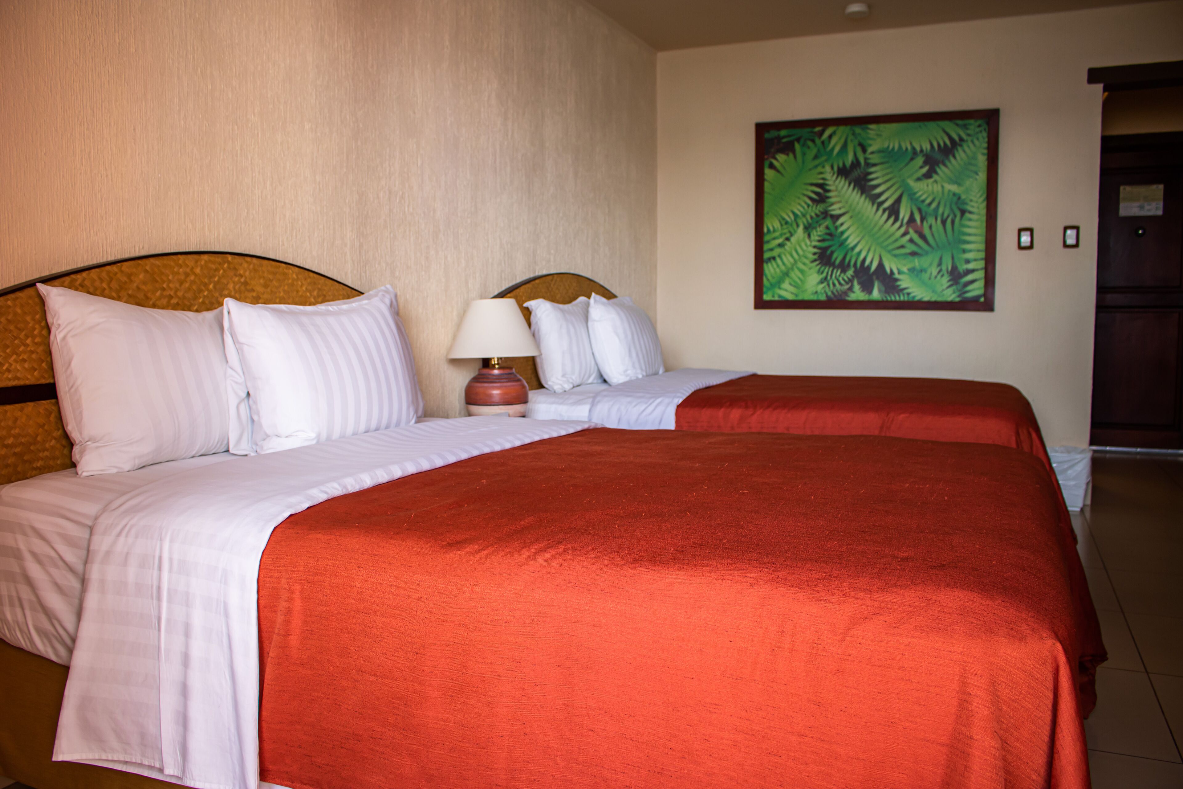 Une chambre d’hôtel avec deux lits, une table de chevet, une lampe et un tableau représentant une feuille tropicale accroché au mur.