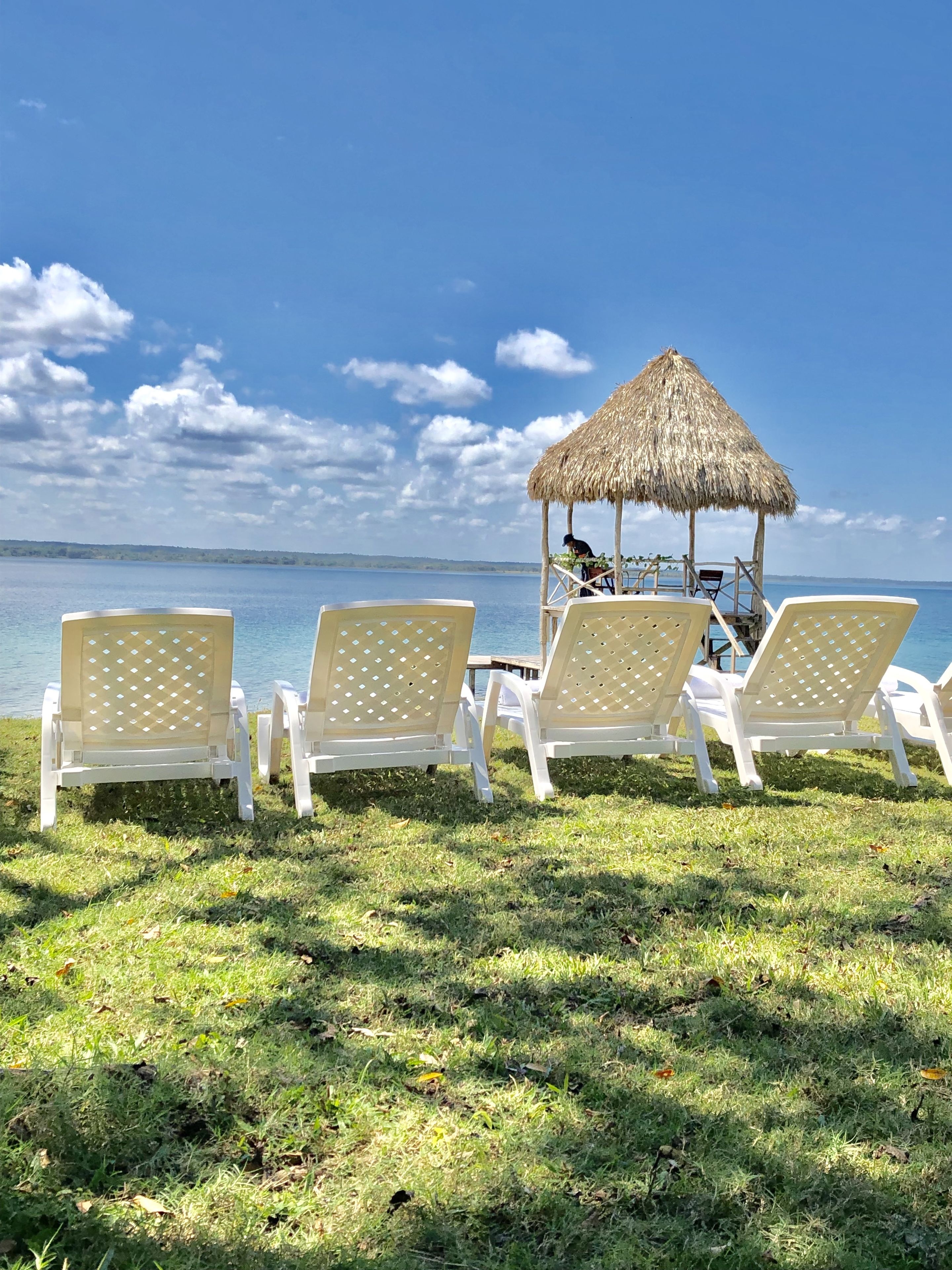 Um espaço de relaxamento à beira-mar, com espreguiçadeiras brancas e uma estrutura com telhado de palha.