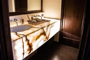 A bathroom with a marble sink, a wooden basket, and a dark wooden door. - Etnea suite 668 (Catania)