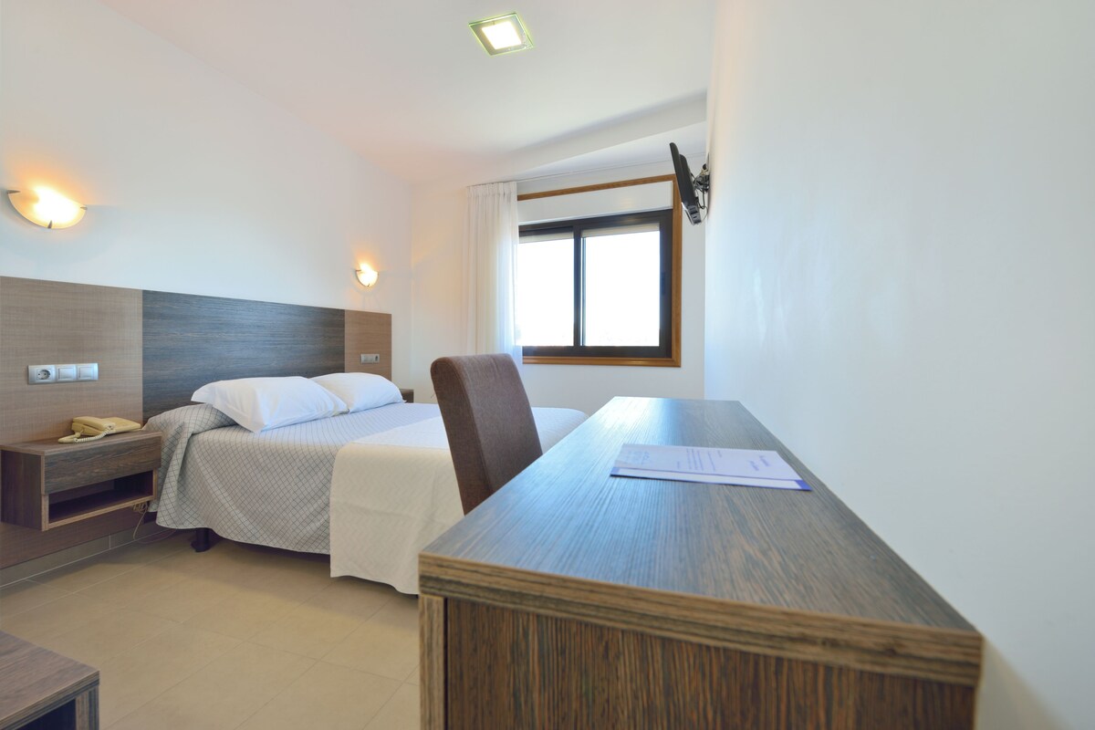 Une chambre d’hôtel avec un lit, un bureau, une chaise, une table de chevet et une fenêtre avec des rideaux.