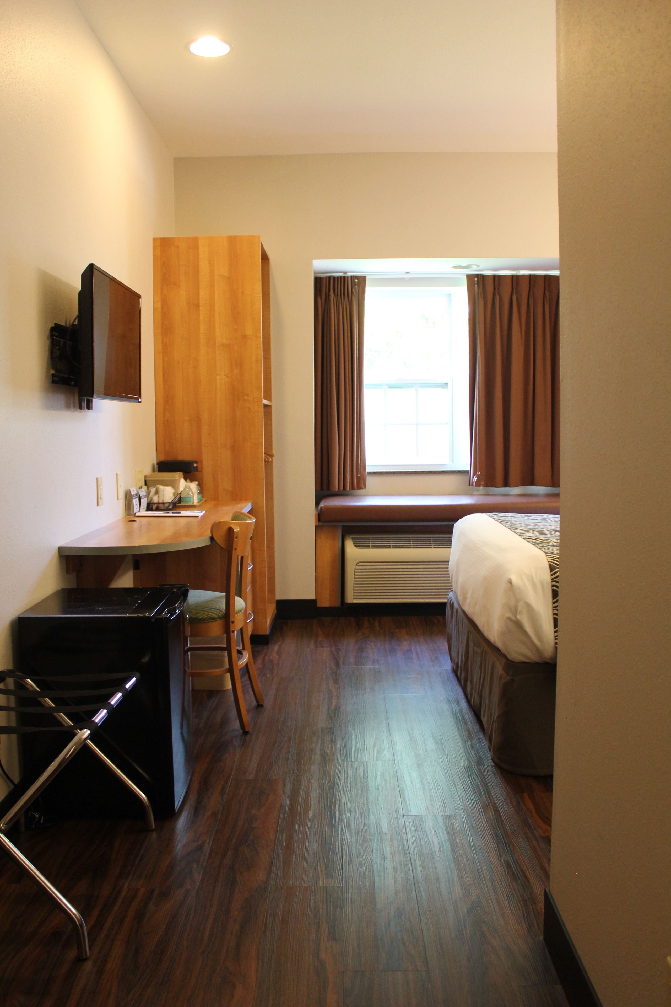 A compact hotel room with a kitchenette, a bed, a window with curtains, and a built-in heater.