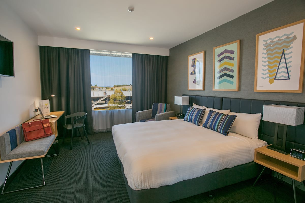 Ein Hotelzimmer mit einem großen Bett, einem Schreibtisch mit einem Laptop, einem Sessel, einem Nachttisch mit Lampe und einem Fenster mit Vorhängen.