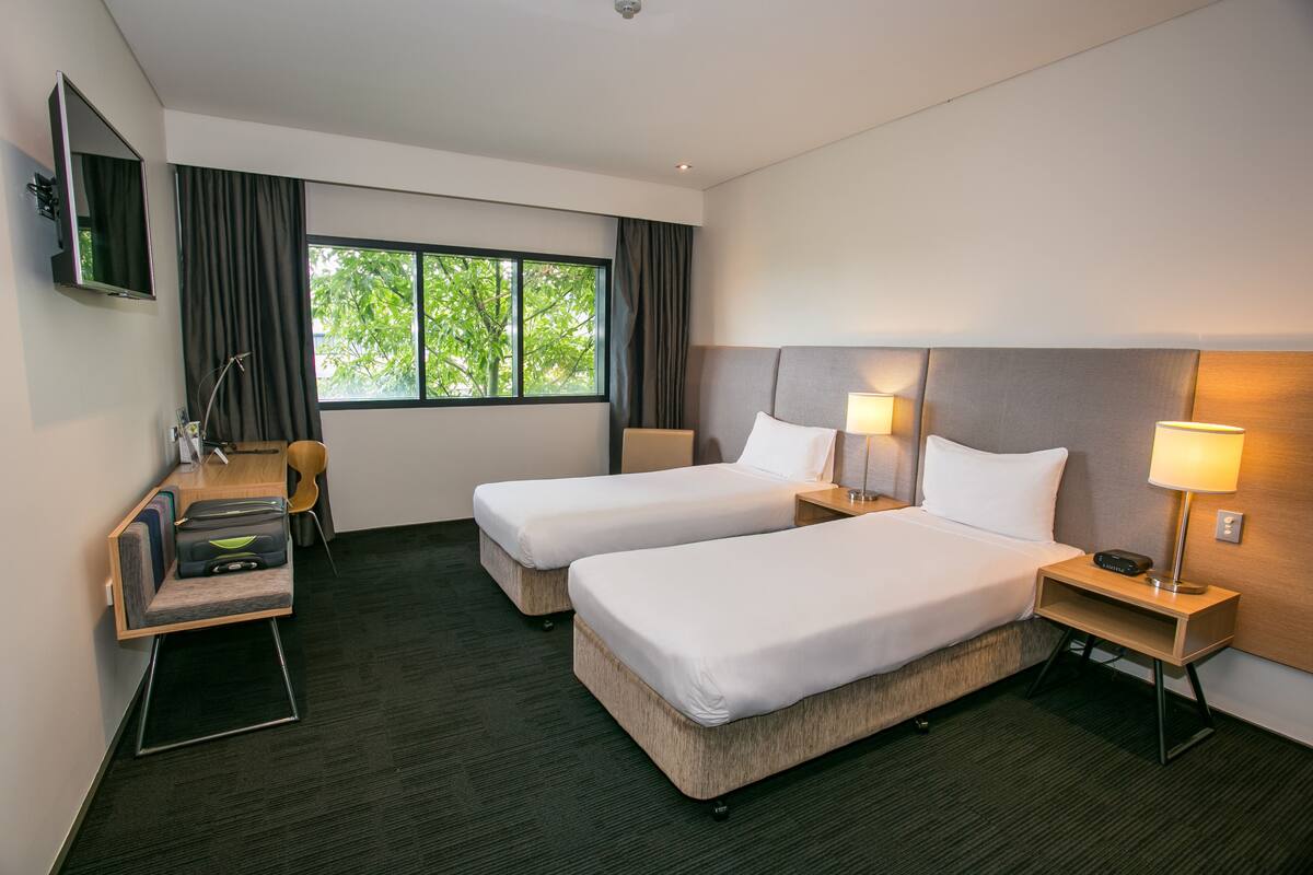 Ein Hotelzimmer mit zwei Betten, einem Schreibtisch, einem Stuhl, einem Fernseher und einem Fenster mit Vorhängen.