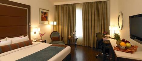 A hotel room with a large bed, a desk with a computer, a chair, a small table with a lamp, and a fruit bowl.