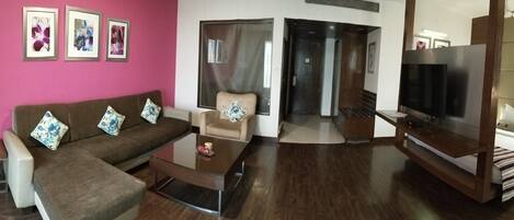 A living room with a brown sofa, a glass coffee table, a TV, and a bed with a striped blanket.