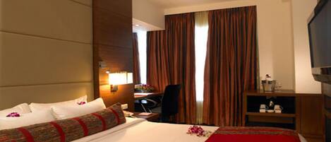 Habitación de hotel con una cama grande, un escritorio, una silla, un televisor y una ventana con cortinas.
