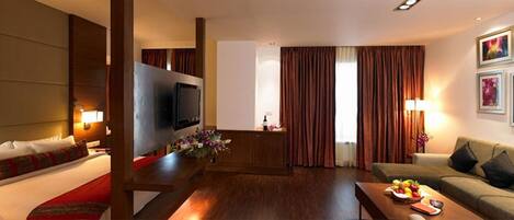 A hotel room with a TV, a sofa, a dining table with fruits, and a flower arrangement.