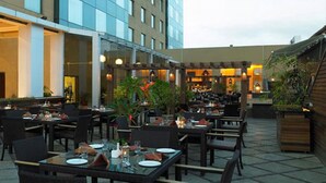 An outdoor dining area with tables and chairs, a covered section with seating, and a building with large windows in the background. - Royal Orchid Central Pune (Pune)