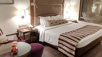 A hotel room with a bed, a nightstand, a chair, a small table, and a glass table with an apple and a jar.